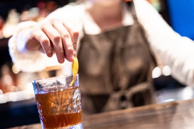 A bartender wearing an apron garnishes a glass of cocktail with a strip of citrus peel at a bar, focusing on the hand and drink. The filename suggests a beverage preparation scene.