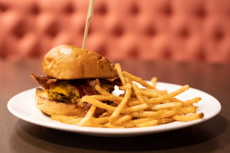 Cheeseburger with bacon and French fries on a white plate, placed on a table with a blurred booth background. The filename suggests focus on burgers.