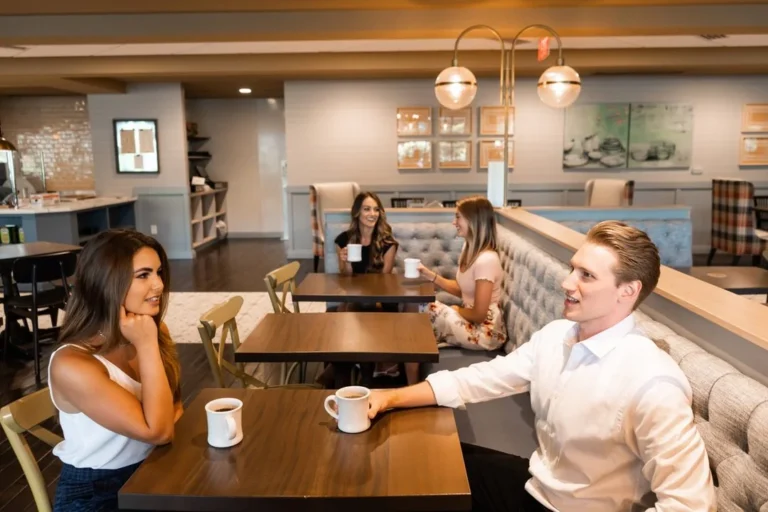 Four young adults sit at tables in a modern cafe, talking and holding coffee mugs. The setting appears relaxed and social, emphasizing conversation and community.