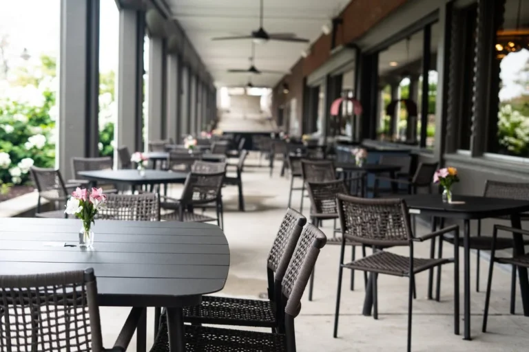 Empty outdoor restaurant patio with black tables and woven chairs, each table featuring a small vase of flowers. The covered area is lined with large windows and fans, suggesting a casual dining environment.