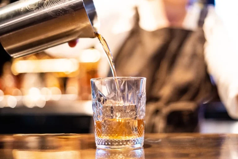A bartender pours whiskey from a shaker into a crystal glass with ice at a bar. Focus on the drink preparation, suggesting a cocktail or neat spirit being served.