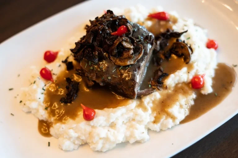 Plated dish featuring braised beef topped with mushrooms, served over mashed potatoes with brown gravy and small red garnish; upscale restaurant presentation.