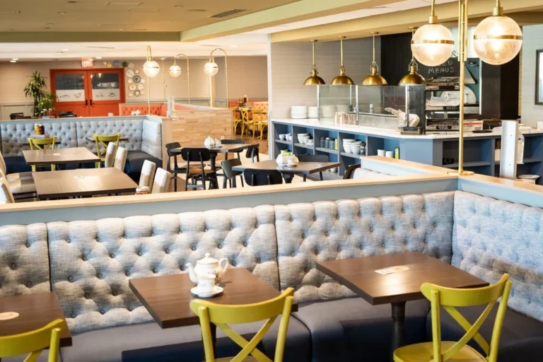 A brightly lit, modern restaurant dining area with light blue tufted booth seating, wooden tables, yellow chairs, and an open kitchen counter with brass pendant lights. The space appears clean and inviting.