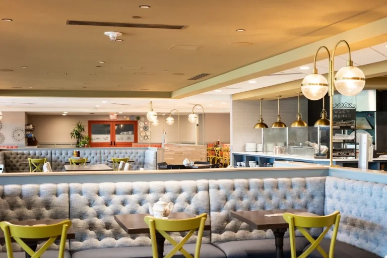Spacious, modern restaurant interior with tufted blue booths, yellow chairs, and pendant lighting. The area is clean and empty, suggesting it is ready for guests. Open kitchen and dining tables visible in the background.