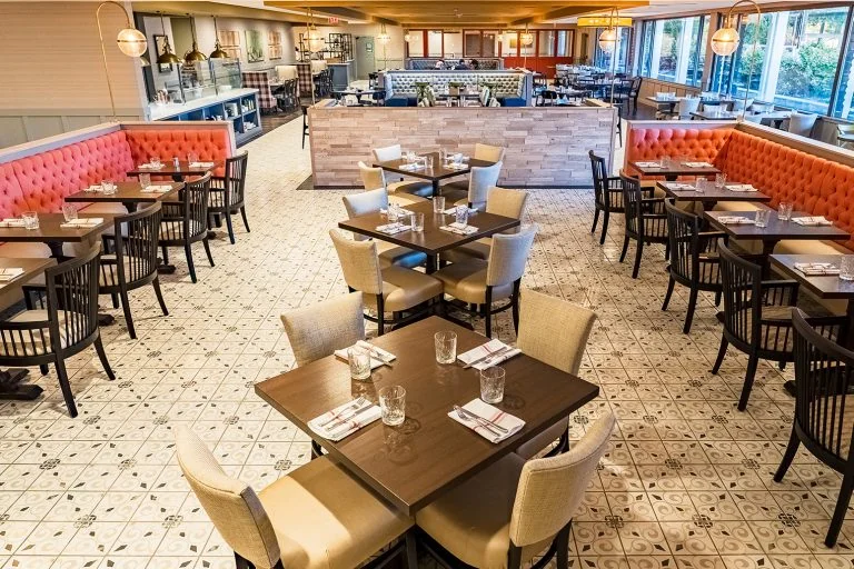Spacious, modern restaurant interior with tufted blue booths, yellow chairs, and pendant lighting. The area is clean and empty, suggesting it is ready for guests. Open kitchen and dining tables visible in the background.