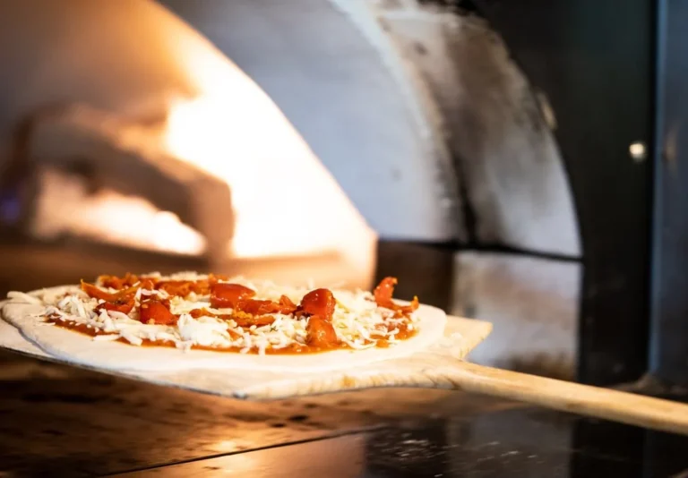 A pizza baking on the stone surface of a wood-fired oven, with visible flames and a rustic setting, captured in a close-up shot.