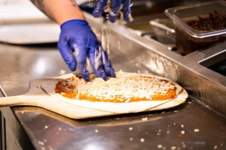 A person wearing blue gloves sprinkles shredded cheese over pizza dough with sauce, preparing pizza in a professional kitchen setting.
