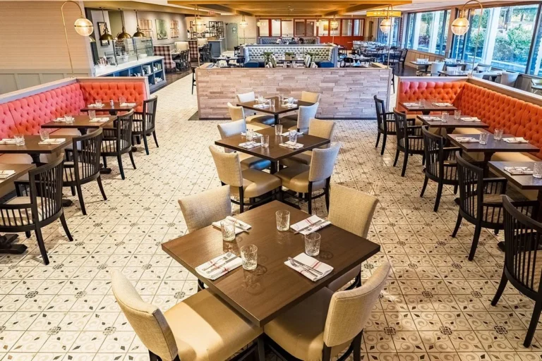 Spacious, well-lit restaurant interior with modern decor, orange tufted booth seating, tiled floors, and neatly set tables. Large windows line one side, providing natural light. The setting suggests a clean and inviting dining environment.